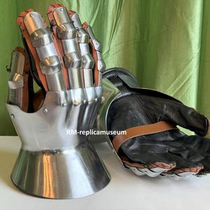 May include: A pair of medieval gauntlets. The gauntlets feature a polished metal exterior with articulated finger plates and a leather interior. The gloves have a black leather palm and a brown leather strap.