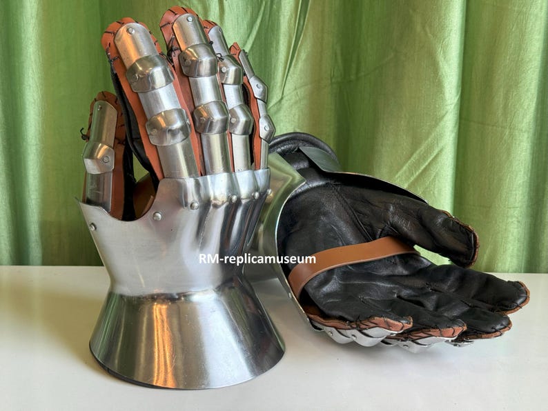 May include: A pair of medieval gauntlets. The gauntlets feature a polished metal exterior with articulated finger plates and a leather interior. The gloves have a black leather palm and a brown leather strap.