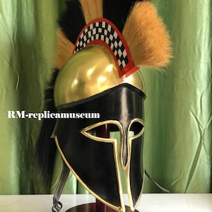 May include: A replica Roman helmet with a gold-colored top and black face guard. The helmet features a black and tan plume and a red and white checkered accent. The text "RM-replicamuseum" is visible on the helmet.