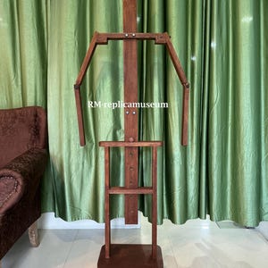 May include: A wooden artist's mannequin with articulated arms and legs. The mannequin is made of dark brown wood and has a rectangular base. The text "RM-replicamuseum" is visible on the mannequin.
