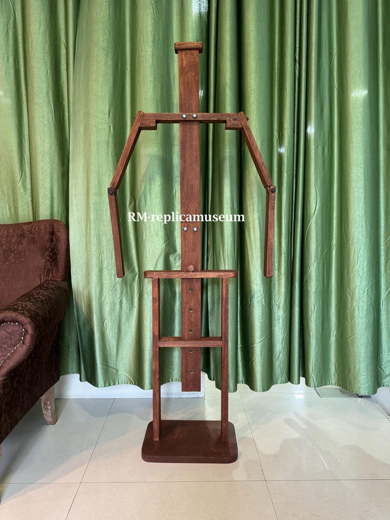 May include: A wooden artist's mannequin with articulated arms and legs. The mannequin is made of dark brown wood and has a rectangular base. The text "RM-replicamuseum" is visible on the mannequin.