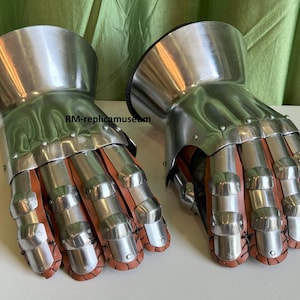 May include: A pair of medieval-style gauntlets. The gauntlets are made of polished metal and feature articulated finger plates and a flared cuff. The interior is lined with brown leather. The gauntlets are designed to protect the hands and wrists.