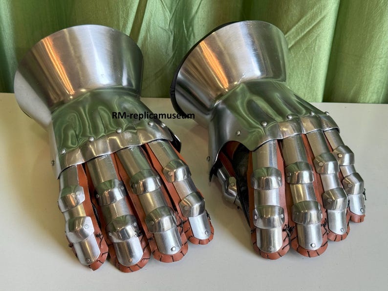 May include: A pair of medieval-style gauntlets. The gauntlets are made of polished metal and feature articulated finger plates and a flared cuff. The interior is lined with brown leather. The gauntlets are designed to protect the hands and wrists.
