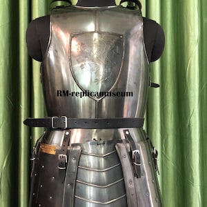 May include: A polished steel breastplate and tasset armor set, displayed on a black mannequin. The breastplate features a shield with a lion emblem. The armor includes a black leather belt and straps with buckles. The mannequin has a size 42 label.