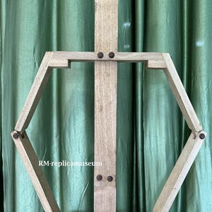 May include: A wooden artist's easel with a light brown finish. The easel features a central vertical support and an adjustable hexagonal frame. The text "RM-replicamuseum" is visible on the frame.