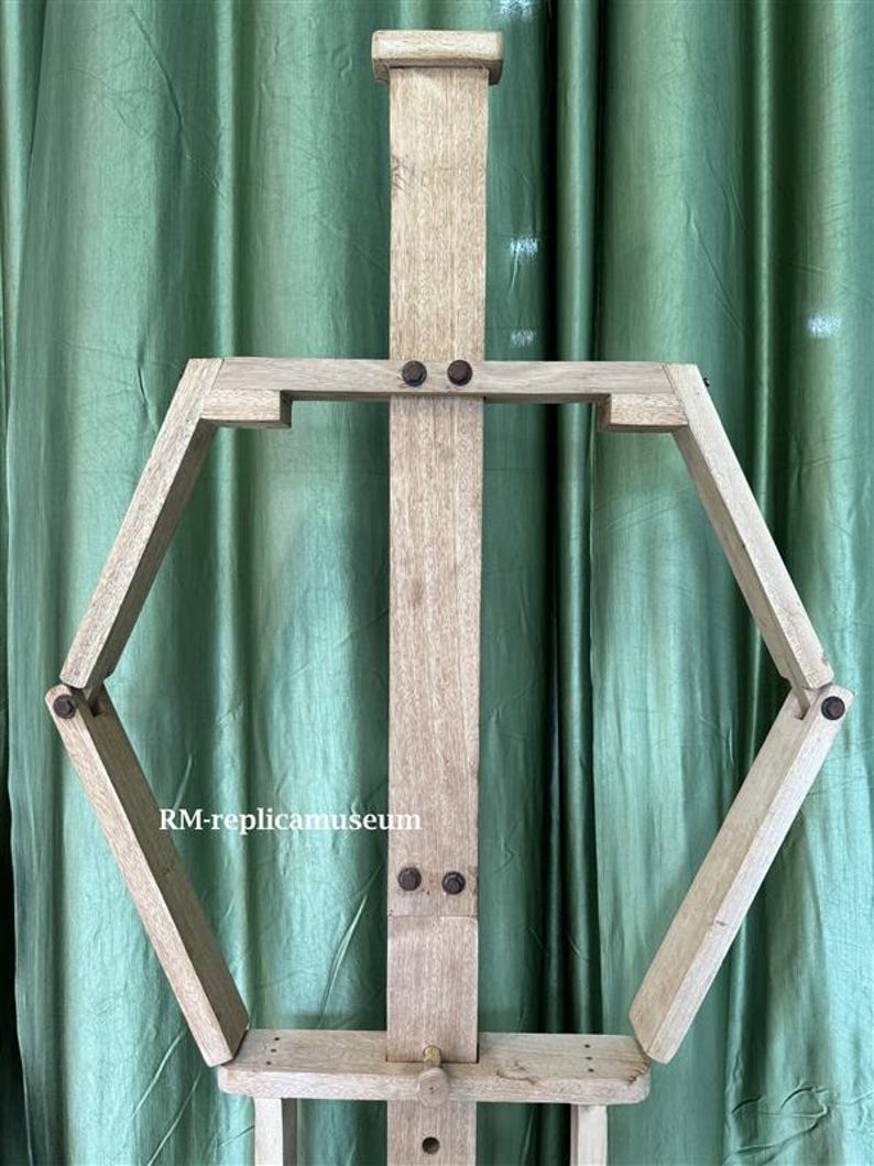 May include: A wooden artist's easel with a light brown finish. The easel features a central vertical support and an adjustable hexagonal frame. The text "RM-replicamuseum" is visible on the frame.