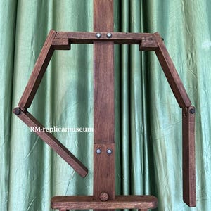 May include: A dark brown wooden artist's easel with adjustable arms and a lower shelf. The easel has a vertical support beam and is designed to hold a canvas for painting. The text "RM-replicamuseum" is visible on the side.