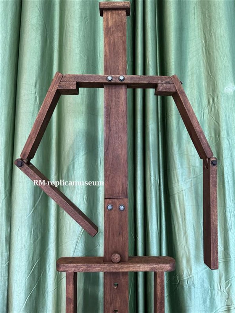 May include: A dark brown wooden artist's easel with adjustable arms and a lower shelf. The easel has a vertical support beam and is designed to hold a canvas for painting. The text "RM-replicamuseum" is visible on the side.