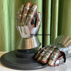 May include: Two medieval gauntlets, one upright on a black stand and the other lying down. The gauntlets are made of polished steel and brown leather, with articulated fingers and a flared cuff. The background is a green curtain.