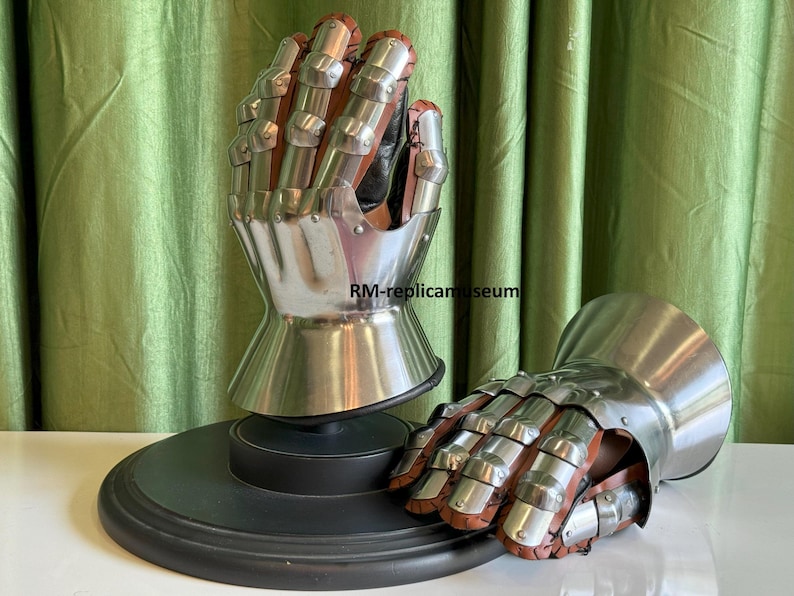 May include: Two medieval gauntlets, one upright on a black stand and the other lying down. The gauntlets are made of polished steel and brown leather, with articulated fingers and a flared cuff. The background is a green curtain.