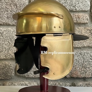 May include: A brass Roman helmet replica, displayed on a wooden stand. The helmet features a rounded top, a visor, and cheek guards. The text "RM-replicamuseum" is visible on the cheek guard.
