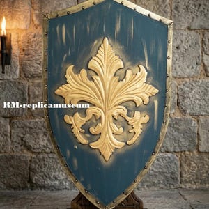 Può includere: Uno scudo decorativo con sfondo blu e design a giglio dorato. Lo scudo ha un bordo dorato ed è montato su un supporto in legno. Il testo "RM-replicamuseum" è visibile.