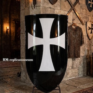 Può includere: Uno scudo nero con una croce bianca, esposto su un supporto metallico. Lo scudo è in una stanza con pareti in pietra, illuminata da applique e un tappeto. Altri scudi e spade sono visibili sullo sfondo.