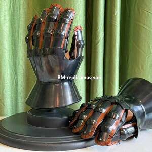 May include: A pair of black and brown medieval gauntlets. The gauntlets are made of metal and leather, with articulated fingers and a flared cuff. One gauntlet is mounted on a black circular base. The other gauntlet is lying on a white surface.