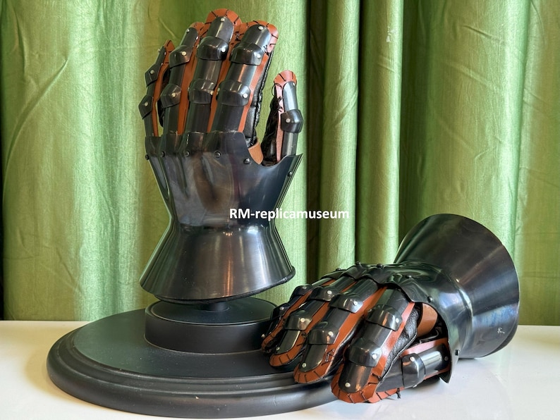 May include: A pair of black and brown medieval gauntlets. The gauntlets are made of metal and leather, with articulated fingers and a flared cuff. One gauntlet is mounted on a black circular base. The other gauntlet is lying on a white surface.