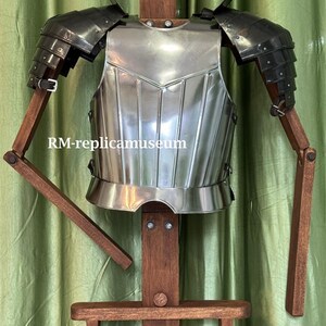 May include: A polished steel breastplate and shoulder armor, displayed on a wooden stand. The armor has a reflective surface and is designed with layered plates. The text "RM-replicamuseum" is visible on the image. The background is a green curtain.