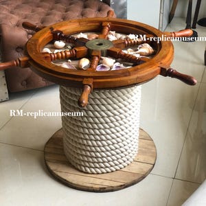 Op de afbeelding: Een koffietafel met nautisch thema, met een houten scheepsrad als bovenkant, gevuld met schelpen. De tafel staat op een cilindrische basis omwikkeld met dik, crèmekleurig touw en een houten basis. Het ontwerp roept een kust- of maritieme esthetiek op.
