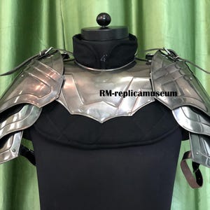 Könnte beinhalten: Ein silberfarbenes Schulterpanzer-Set aus Metall mit geschichteten Platten und Lederriemen. Die Rüstung wird auf einer schwarzen Schaufensterpuppe mit schwarzem Kragen und grünem Vorhanghintergrund präsentiert. Der Text "RM-replicamuseum" ist sichtbar.