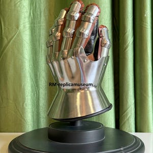 May include: A detailed replica of a medieval gauntlet, featuring articulated metal plates and brown leather accents. The gauntlet is mounted on a black display stand, showcasing its intricate design and craftsmanship. The text "RM-replicamuseum" is visible.