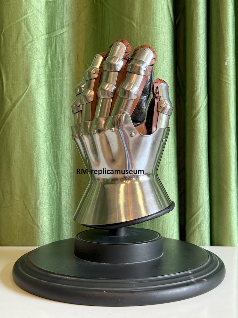 May include: A detailed replica of a medieval gauntlet, featuring articulated metal plates and brown leather accents. The gauntlet is mounted on a black display stand, showcasing its intricate design and craftsmanship. The text "RM-replicamuseum" is visible.