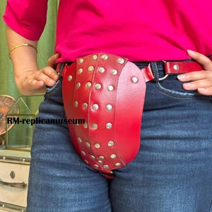 May include: A red leather garment with silver studs, worn over blue jeans and a red belt. The garment has a curved shape and is secured with a belt. The person is wearing a pink shirt.