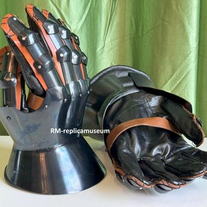 May include: Pair of black and orange medieval gauntlets. The gauntlets feature articulated finger plates and a flared cuff. The leather gloves are secured with brown straps. The armor is designed for protection and historical reenactment.