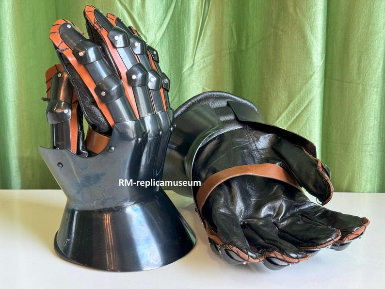 May include: Pair of black and orange medieval gauntlets. The gauntlets feature articulated finger plates and a flared cuff. The leather gloves are secured with brown straps. The armor is designed for protection and historical reenactment.