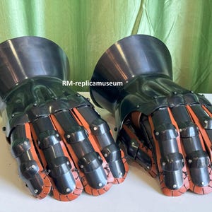May include: Pair of black and orange articulated metal gauntlets. The gauntlets have a polished finish and are designed to protect the hands. The fingers are segmented with orange leather or fabric visible between the metal plates. The gauntlets are likely for historical reenactment or display.