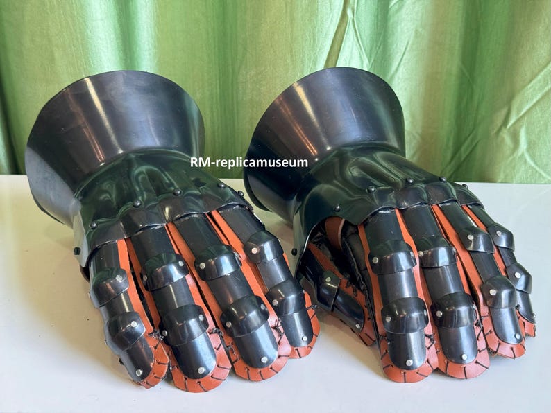 May include: Pair of black and orange articulated metal gauntlets. The gauntlets have a polished finish and are designed to protect the hands. The fingers are segmented with orange leather or fabric visible between the metal plates. The gauntlets are likely for historical reenactment or display.