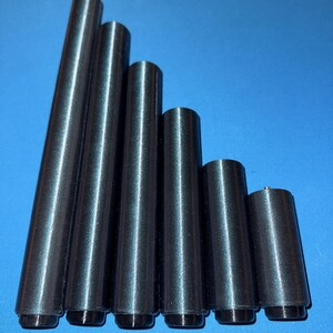 Può includere: Sei oggetti cilindrici in metallo di diverse altezze, disposti in fila. Gli oggetti sono grigio scuro con una superficie riflettente e una base nera. Lo sfondo è blu unito.