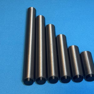 Può includere: Sei oggetti cilindrici in metallo di varie lunghezze, disposti in ordine decrescente. I cilindri sono grigio scuro con tappi neri sul fondo. Lo sfondo è blu chiaro uniforme.