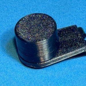 May include: A black, glitter-covered money clip with a cylindrical top. The clip is made of a dark material and sits on a blue surface. The cylindrical top has a textured, sparkling appearance.