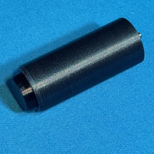 Può includere: Un oggetto cilindrico nero con una base piatta e circolare e una piccola punta argentata. L'oggetto ha una superficie strutturata e sembra essere fatto di un materiale solido. È appoggiato su una superficie blu.
