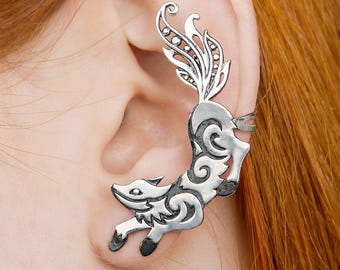 May include: A silver-colored ear cuff in the shape of a fox. The fox is leaping, with intricate scrollwork detailing its body and a decorative tail. The ear cuff is designed to wrap around the ear.