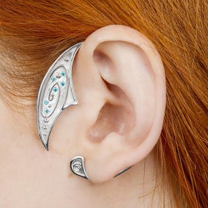 May include: Silver ear cuff with an elven design, featuring a curved shape that wraps around the ear. The cuff has a series of small, light blue gemstones and decorative detailing. The design includes a smaller piece for the earlobe.