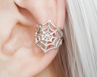 Puede incluir: Puño de oreja plateado con diseño de telaraña. El puño se lleva en la oreja y tiene un patrón detallado y calado. Una joya de estilo gótico o con temática de Halloween.