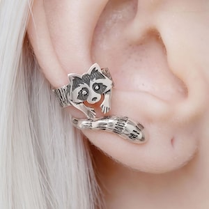 Puede incluir: Un pendiente de oreja plateado con un diseño de mapache. El pendiente se envuelve alrededor de la oreja, con la cabeza del mapache en la parte superior y su cola rayada extendiéndose hacia abajo. El mapache tiene rasgos faciales detallados.