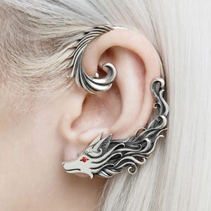 May include: A silver ear cuff with a wolf design and red eyes. The cuff wraps around the ear with detailed, flowing lines and a stylized wolf head. The jewellery is worn on the ear.