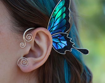 May include: A butterfly ear cuff with a silver wire design and a large, iridescent blue and green wing. The wing has a black glitter outline. The ear cuff wraps around the ear and features two spiral designs.
