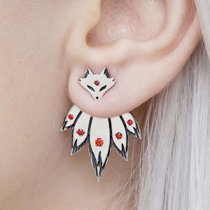 Puede incluir: Pendientes de zorro plateados con detalles de gemas rojas. El perno superior es una cabeza de zorro, y la pieza trasera presenta un diseño en forma de abanico con gemas rojas. Los pendientes se lucen en una oreja.