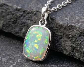 May include: A rectangular pendant featuring a white opal with iridescent green and yellow flashes, set in a polished silver frame. The pendant hangs from a delicate silver chain, showcasing a smooth surface and a bright, reflective finish.