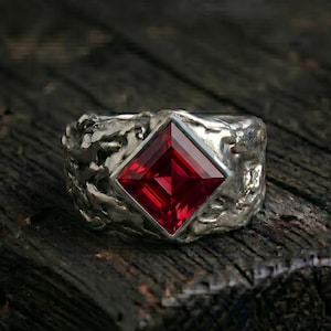 Può includere: Un anello in argento con una pietra preziosa rosso scuro dal taglio quadrato incastonata al centro. La fascia presenta una superficie ruvida, irregolare e testurizzata che contrasta con la superficie liscia e sfaccettata della pietra rossa, creando un aspetto visivo distinto.