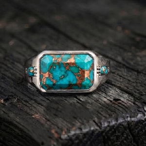 Könnte beinhalten: Ein Silberring mit einem rechteckigen, facettierten Türkisstein mit markanter kupferfarbener Matrix-Maserung. Die Ringschiene ist auf beiden Seiten der zentralen Steinfassung mit kleinen, runden Türkis-Cabochons verziert.