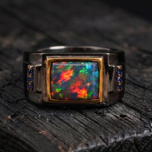 Op de afbeelding: Een ring met een rechthoekige zwarte opaal met levendige rode, groene en blauwe flitsen, gezet in een gouden rand. De ringband is van donker metaal met een matte afwerking, geaccentueerd door kleine blauwe steentjes aan de zijkanten.