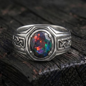 May include: A silver ring featuring an oval-cut dark opal gemstone with vibrant red, orange, and green flashes. The band is crafted from polished silver and displays an intricate, repeating geometric knotwork pattern along both sides of the setting.