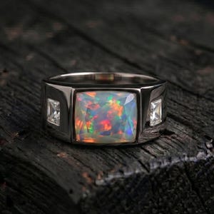 Könnte beinhalten: Ein silberfarbener Ring mit einem großen, rechteckigen, mehrfarbigen Opal in der Mitte. Die Ringschiene ist auf beiden Seiten des zentralen Opals mit zwei kleineren, klaren, quadratisch geschliffenen Steinen in einer polierten Metallfassung besetzt.