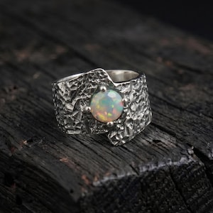 May include: A textured silver ring with a round, iridescent opal gemstone. The ring has a wide band with a rugged, hammered appearance. The opal displays a range of colors, including white, yellow, and hints of blue and green. The ring is set against a dark, wooden surface.