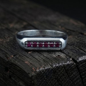 May include: A silver ring with a rectangular face set with six small, vibrant red gemstones. The ring is displayed on a dark, textured wooden surface, highlighting the contrast between the metal and the gemstones.