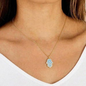 May include: A gold-coloured necklace featuring a Hamsa hand pendant. The pendant has a light blue enamel design with white detailing and a central eye. The necklace is worn against a white vest top.