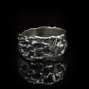 May include: A silver ring with a textured, organic, and uneven surface. The metal has a dark, oxidized finish in the recessed areas, creating a high-contrast look with the polished, raised sections of the band. The ring has a wide, irregular shape.